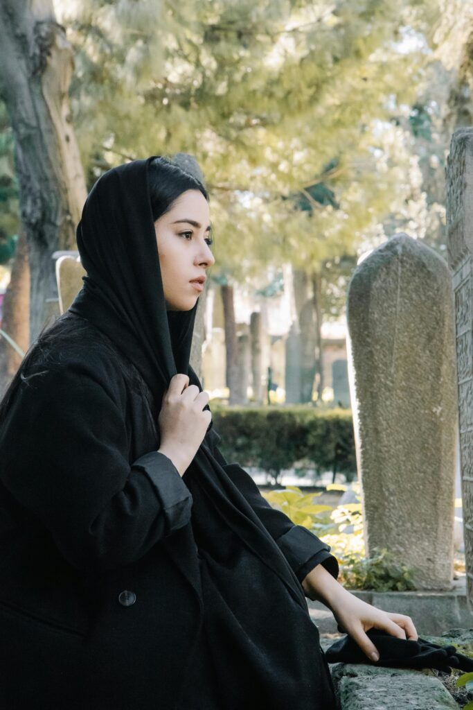 Side view of calm ethnic female wearing traditional black clothes and headwear sitting near aged stone gravestones on obsolete cemetery